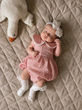 A sleeping newborn baby wearing a pink and white gingham romper, matching headband, and white knit socks, lying on a beige quilted blanket next to a white plush duck toy.