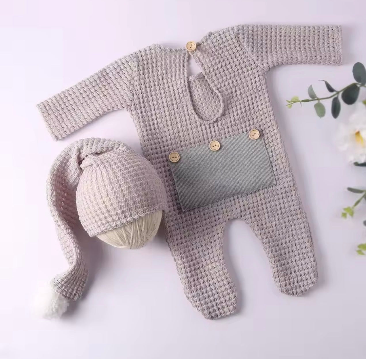 A baby knit outfit set laid flat on a pale pink background, including a long-sleeve footed romper in a textured light beige knit with a gray front patch and wooden buttons, plus a matching knit hat with a long tail and pom-pom. Decorative flowers appear at the edges.

Shopify Tags:
newborn outfit, baby romper, waffle knit romper, newborn romper, baby boy outfit, baby girl outfit, gender neutral baby, newborn coming home, coming home outfit, newborn photo outfit, baby photoshoot outfit, newborn photography, 