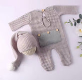 A baby knit outfit set laid flat on a pale pink background, including a long-sleeve footed romper in a textured light beige knit with a gray front patch and wooden buttons, plus a matching knit hat with a long tail and pom-pom. Decorative flowers appear at the edges.

Shopify Tags:
newborn outfit, baby romper, waffle knit romper, newborn romper, baby boy outfit, baby girl outfit, gender neutral baby, newborn coming home, coming home outfit, newborn photo outfit, baby photoshoot outfit, newborn photography, 