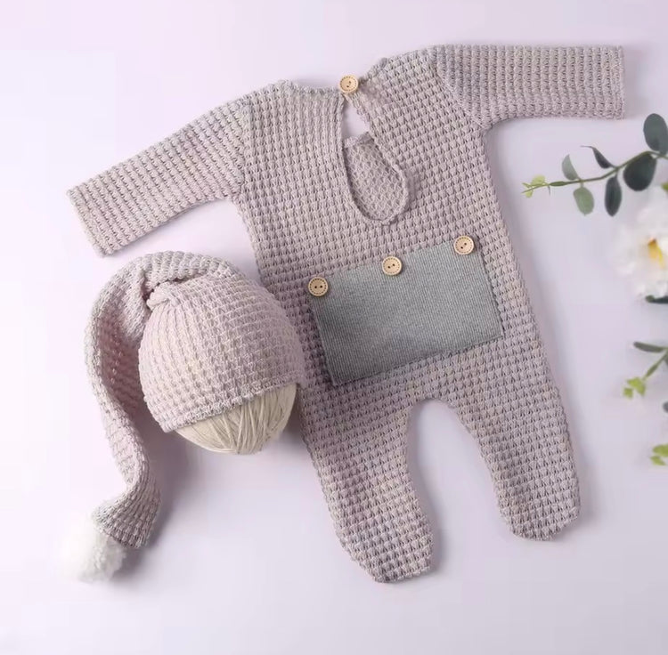 A baby knit outfit set laid flat on a pale pink background, including a long-sleeve footed romper in a textured light beige knit with a gray front patch and wooden buttons, plus a matching knit hat with a long tail and pom-pom. Decorative flowers appear at the edges.

Shopify Tags:
newborn outfit, baby romper, waffle knit romper, newborn romper, baby boy outfit, baby girl outfit, gender neutral baby, newborn coming home, coming home outfit, newborn photo outfit, baby photoshoot outfit, newborn photography, 