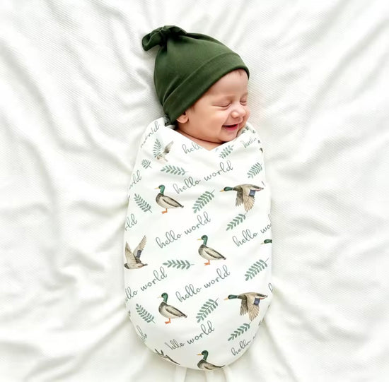 A sleeping newborn baby swaddled in a white muslin swaddle blanket with a green and brown duck print and &