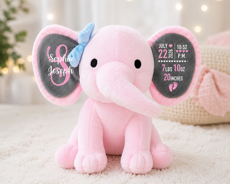 A pink plush elephant toy sitting on a cream carpet in a softly lit room. The elephant has large dark gray ears with custom printed names and birth details, a light blue bow on its left ear, and black eyes.